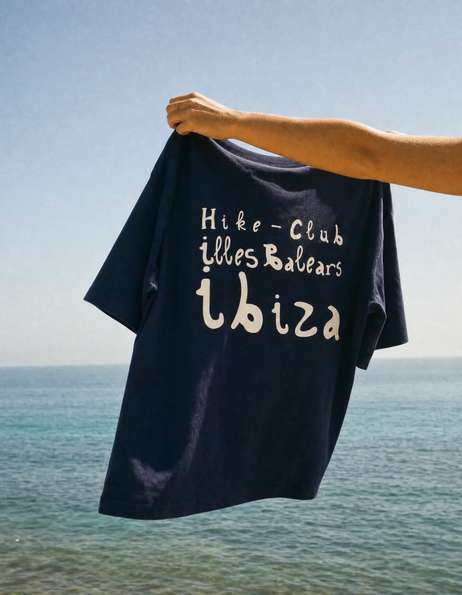Navy Hike Club Ibiza T-shirt held up in front of the sea